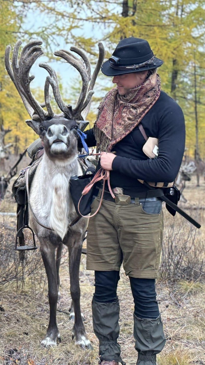 A person standing beside a reindeer with large antlers among autumn trees.
