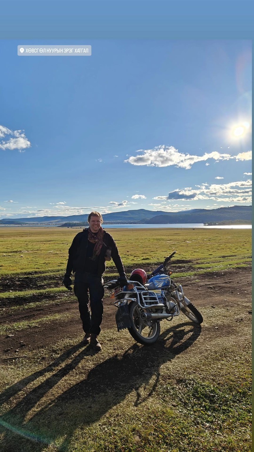 A person standing beside a blue motorcycle near a lake under a bright sky.