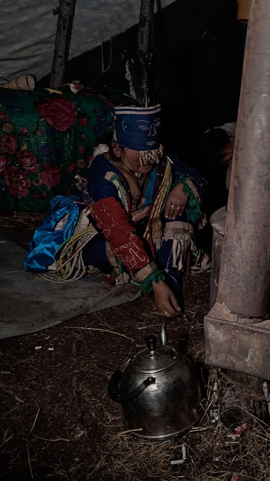 Tsetse seated in layered blue and red ceremonial clothing beside a metal kettle in a dark interior.