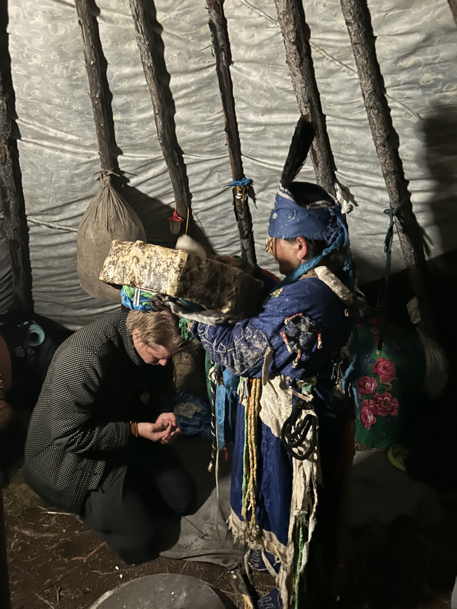 Tsetse holds a large drum over a kneeling person inside a taiga dwelling.